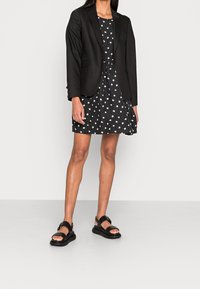 Mustat räätälöidyt bleiserit mustan mekon päällä, jossa on valkoisia pilkkuja; litteät mustat sandaalit, joissa on hihnat ja paksu pohja. Sujuva pinta.