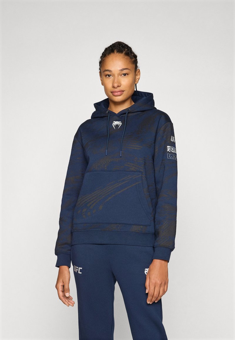 Felpa navy con un motivo sottile, tasca frontale, cappuccio con cordini e dettagli con logo su petto e manica. Il materiale sembra morbido e testurizzato.