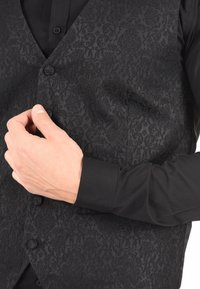 Gilet nero con un motivo testurizzato, sei bottoni e una vestibilità su misura, indossato sopra una camicia nera a maniche lunghe.