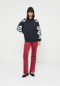 Mujer de pie con la mano en la cadera, vistiendo un sudadera negra con grandes diseños florales blancos y pantalones rojos, contra un fondo blanco.