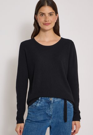 Femme aux longs cheveux foncés portant un haut noir à manches longues et un jean bleu, debout devant un fond clair uni.
