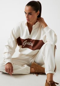 Felpa bianca con zip e una striscia marrone con il logo "Guess"; abbinata a pantaloni jogger bianchi e scarpe marroni con lacci.
