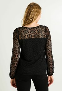 Top de encaje negro con mangas largas y un patrón de ondas, un panel sólido negro en la parte posterior y un escote redondo. Textura semitranslúcida.