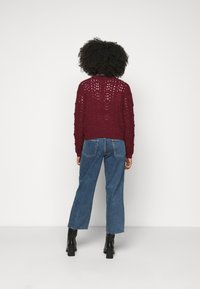 Maglione a maglia bordeaux con design a motivo traforato, scollo rotondo e orlo a costine; abbinato a jeans in denim a vita alta e stivaletti neri.
