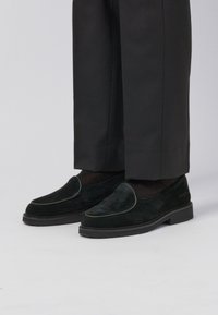Black suede loafers with a smooth texture, featuring a decorative green piping, worn with dark tailored pants. Low-profile rubber sole.