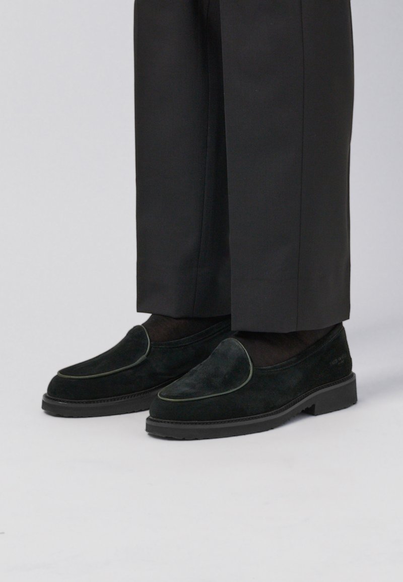 Black suede loafers with a smooth texture, featuring a decorative green piping, worn with dark tailored pants. Low-profile rubber sole.