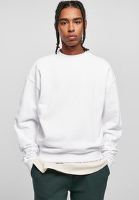 Hvid sweatshirt med løs pasform, dropped skuldre og ribbede manchetter, lagd over en lysefarvet top og parret med mørkegrønne bukser.