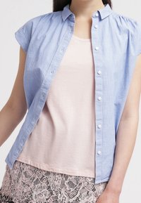 Camicia azzurro chiaro a maniche corte, con bottoni, realizzata in tessuto leggero, indossata sopra un top rosa tenue. Presenta bottoni e colletto.