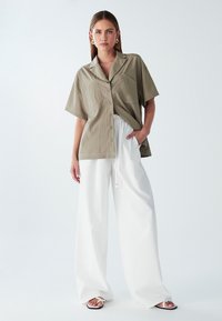 Mujer vestida con una camisa beige de botonadura corta y holgada, pantalones blancos de pierna ancha, sandalias blancas, de pie con una mano en el bolsillo sobre un fondo liso.