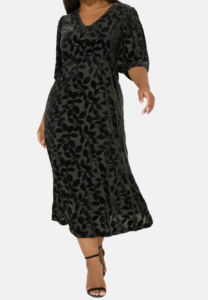 Donna che indossa un vestito midi nero con motivo floreale in velluto, maniche a sbuffo a mezza lunghezza, scollo a V e tacchi alti neri con cinturino alla caviglia.