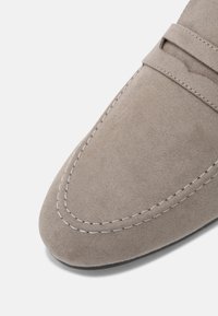 Mocassin en daim gris avec des coutures décoratives, une texture lisse, un bout rond et un design sans lacets comportant une bande sur le dessus.