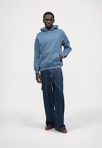 Zila kapuce ar priekšējo kabatu, apvienota ar tumši zilu svītrainu platleğu biksēm un brūnām kurpēm. Vienkāršs, ikdienišķs dizains.