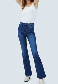 Blaue, ausgestellte Jeans aus Denim mit mittelhohem Bund. Begleitet von einem weißen Tanktop. Glatte Textur, klassisches 5-Taschen-Design, leichte Verwaschung.