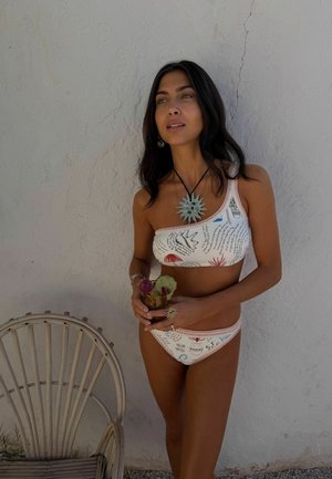 Kobieta w wzorzystym bikini na jedno ramię, trzymająca drinka, stojąca obok drewnianego krzesła na tle teksturowanej białej ściany.