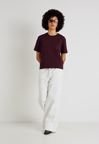 Burgundijas īsām piedurknēm t-krekls apvienots ar baltām plašām biksēm un melniem papēžiem. Modelis valkā saulesbrilles un stāv uz vienkārša fona.