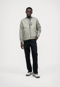 Chaqueta bomber gris claro con cremallera frontal, cuello acanalado y bolsillos laterales, combinada con pantalones oscuros y zapatillas grises. Textura sintética suave.