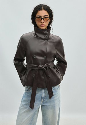 Femme portant une veste en cuir marron foncé ceinturée avec un col montant et un jean bleu clair, debout les mains dans les poches devant un fond uni.