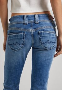 Person in blauen Jeans mit gesteppten Doppel-Taschen-Details und einem weißen, cropped Top, von hinten auf Taillenhöhe gezeigt.