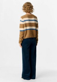 Person mit lockigem, kurzem Haar steht mit dem Rücken zur Kamera, trägt einen braunen, flauschigen Pullover mit blauen und cremefarbenen Streifen sowie dunkelblauen, weiten Hosen.