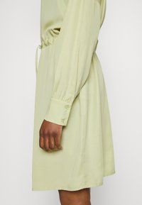 Vestido de manga curta, verde claro, feito de tecido suave. Apresenta uma cintura com cordão e punhos com botões. A vista lateral mostra uma silhueta relaxada.