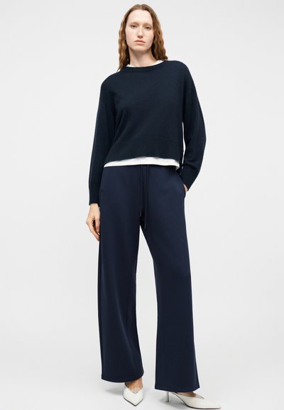 Γυναίκα που φοράει μαύρο πουλόβερ πάνω από λευκό πουκάμισο, φαρδιά μπλε παντελόνια και λευκά μυτερά τακούνια, στέκεται μπροστά σε μια απλή ανοιχτόχρωμη επιφάνεια.