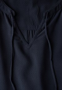 Top blu scuro con scollo a V, dettagli con lacci e un tessuto testurizzato con piccole perforazioni. Dettaglio plissettato allo scollo.