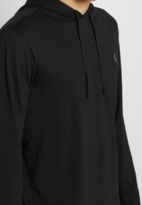 En person som bär en svart långärmad hoodie med dragsko och en liten logotyp på bröstet, stående mot en enfärgad bakgrund.