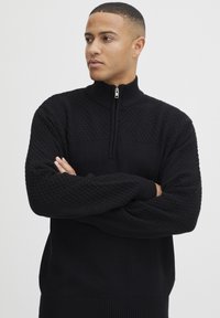 Junger Mann mit kurzen Haaren, der einen schwarzen, strukturierten Strickpullover mit einem Viertel-Reißverschluss trägt, die Arme verschränkt, und zur Seite blickt, vor einem schlichten Hintergrund.