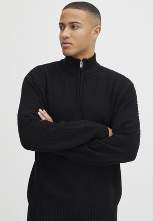 Ung man med kort hår iklädd en svart texturerad stickad tröja med en fjärdedelsdragkedja, med armarna i kors, tittar åt sidan mot en enfärgad bakgrund.