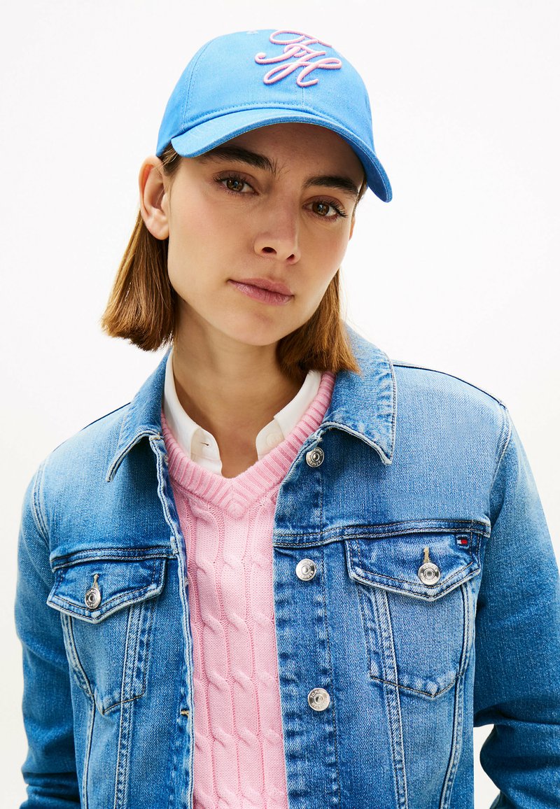 Joven con cabello corto y castaño, lleva una gorra azul, chaqueta de mezclilla azul claro, suéter de punto rosa y camisa blanca sobre un fondo liso.