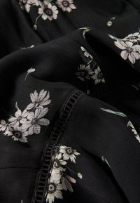 Svart tyg med vitt blommönster som visar olika blommor och gröna blad. Har delikata detaljer längs kanterna. Mjuk textur.