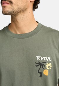 Groene katoenen T-shirt met ronde hals, voorzien van een geborduurd RVCA-logo en abstracte ontwerpen van een palmboom, zon en gezicht in zwart en geel.