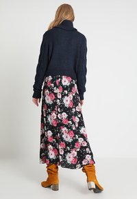 Tumma laivastonsininen neulepusero, jossa on korkea kaulus, pitkän mustan kukkamekon päällä, jossa on vaaleanpunaisia ja valkoisia kukkia. Ruskeat, löysät saapikkaat, joissa on korot.