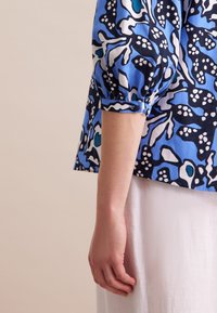 Blaue gemusterte Bluse mit Puffärmeln, die abstrakte Formen in Dunkelblau, Weiß und Türkis zeigt. Der Stoff scheint leicht und strukturiert zu sein.