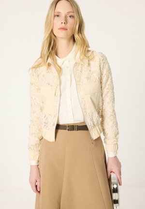 Hõbedast ümmargust sidurit hoidev heledate juustega naine, kes kannab valge nööpidega särkjakiga kreemjat lillelist pitsjopet, pruunikasbeeži seelikut ja pruuni vööd.