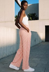 Young woman with short hair stands outdoors wearing a white tank top, pink and white checkered wide-leg pants, and white sneakers.