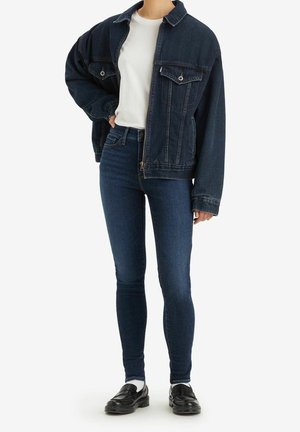 Femme portant une veste en jean bleu foncé, un t-shirt blanc, un jean slim foncé, des chaussettes blanches et des mocassins noirs, debout avec la main dans la poche.