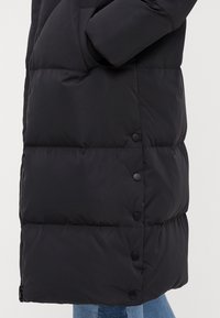 Čierna puffer bunda s hladkou textúrou, so zipsovým zapínaním a čiernymi patentkami pozdĺž spodného okraja. Polopriliehavý, po kolená siahajúci strih.