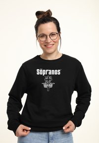 Felpa nera con il testo "The Sopranos" in bianco sopra un grafico di una corona e una croce. Il tessuto appare texturizzato e morbido.