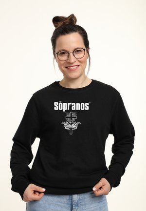 Sort sweatshirt med "The Sopranos" tekst i hvid over et grafisk motiv af en krone og et kors. Stoffet ser tekstureret og blødt ud.