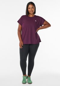 Frau steht mit der Hand auf der Hüfte, trägt ein lila kurzärmliges Shirt, schwarze Leggings und grüne Sneakers vor einem weißen Hintergrund.