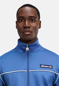 Blaue Trackjacke aus glattem, leichtem Stoff. Merkmale einen hohen Kragen, einen durchgehenden Reißverschluss und kontrastierende weiße Paspelierung. Zeigt ein Logo auf der Brust.