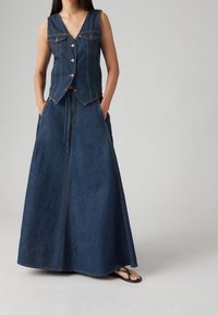 Džínová vesta s výstřihem do V a pěti knoflíky, spárovaná s dlouhou, splývavou džínovou sukní se bočními kapsami a elastickým pasem na šňůrku.