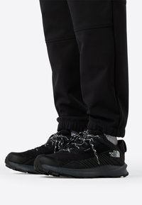 Μαύρα αθλητικά παπούτσια με ανάγλυφο πλέγμα στην επάνω επιφάνεια, κορδόνια με ασπρόμαυρο σχέδιο και γκρι λεπτομέρειες στη σόλα και τη σήμανση.