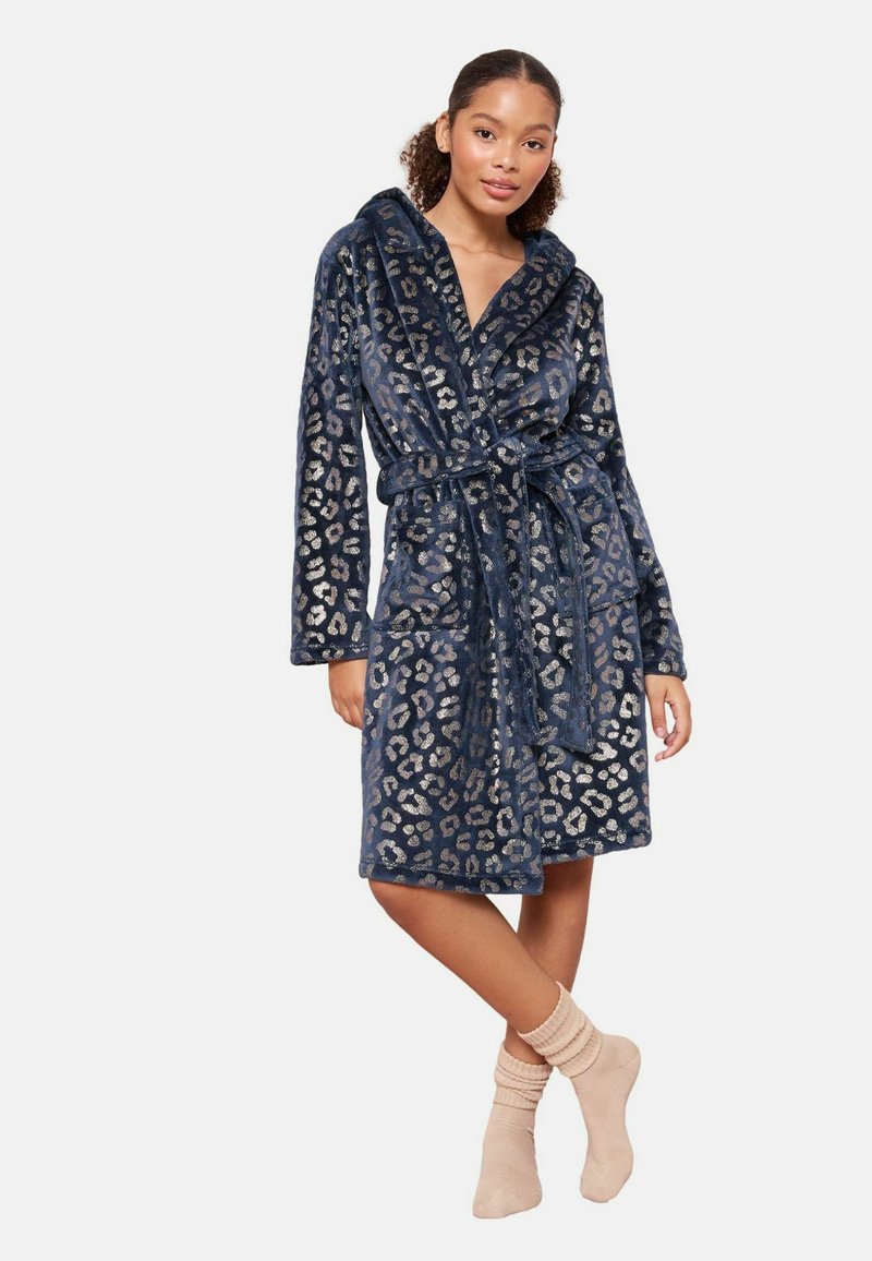 Accappatoio blu navy in peluche con stampa leopardata argentata, design avvolgente, cintura annodata e tasche frontali. Il modello indossa calzini a coste beige con rouches.