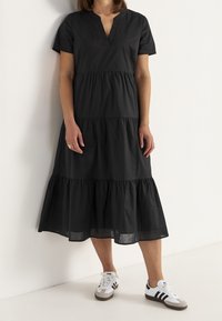 Schwarzes Stufenkleid mit V-Ausschnitt, kurzen Ärmeln und einem fließenden Saum, kombiniert mit weißen Sneakern mit drei schwarzen Streifen.