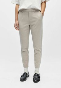 Personne portant un pantalon beige ajusté, des chaussettes blanches, des mocassins noirs, une chemise blanche rentrée et un bracelet argenté au poignet gauche.