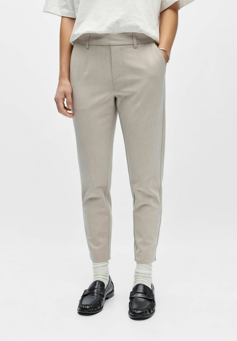 Personne portant un pantalon beige ajusté, des chaussettes blanches, des mocassins noirs, une chemise blanche rentrée et un bracelet argenté au poignet gauche.