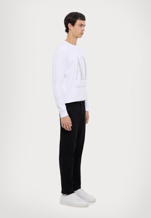 Cămașă albă cu mâneci lungi, cu un logo imprimat, asociată cu pantaloni negri și adidași albi. Design simplu, material din bumbac, linii curate.