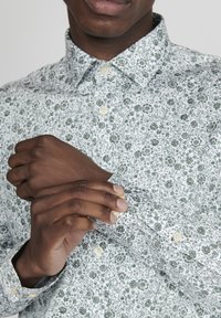 Vit skjorta med knappar och ett grönt blommönster. Har detaljer på krage och manschetter. Tyget verkar slätt och lätt.